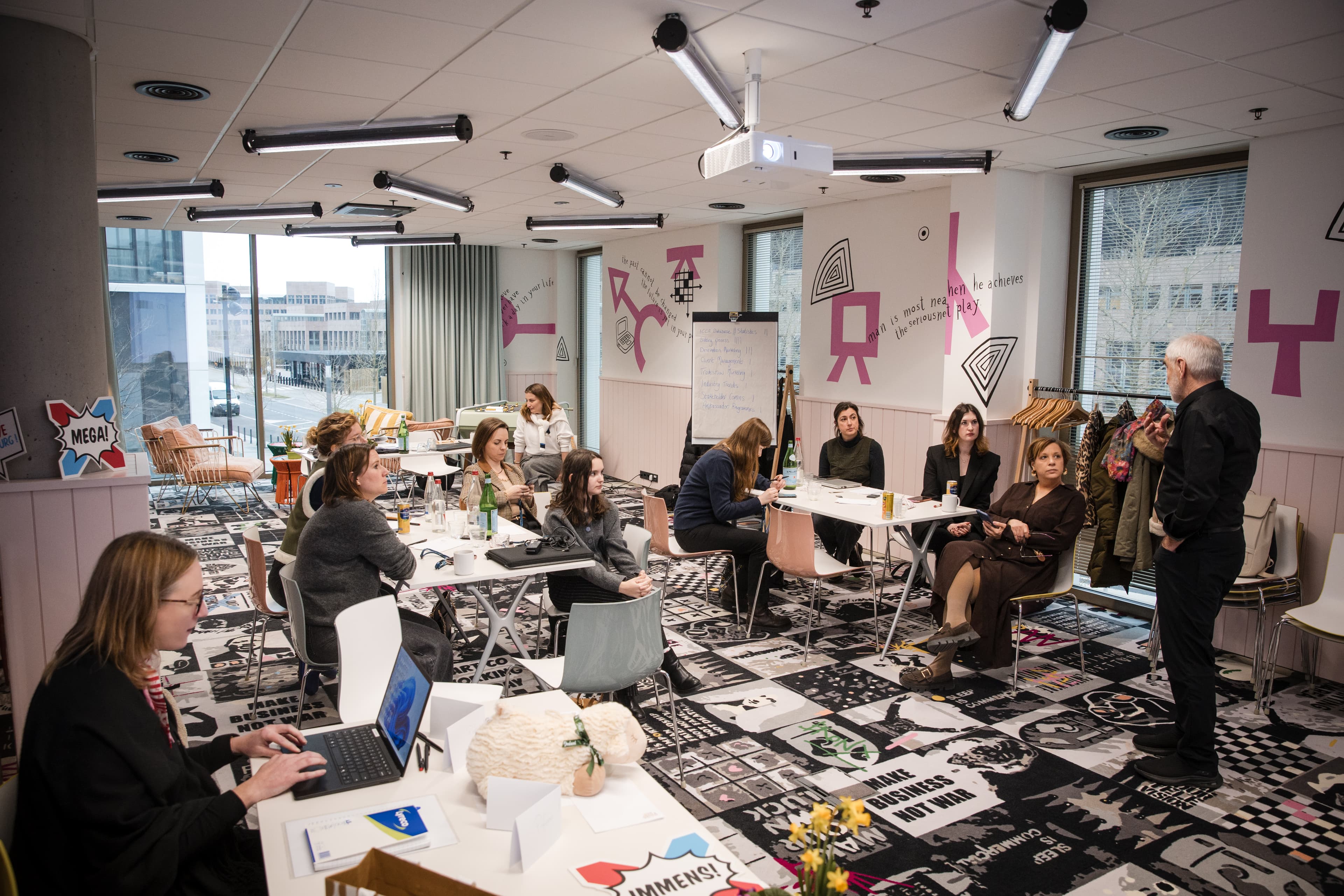 Modern meeting room with participants listening to a presenter, colorful decor and contemporary furniture.