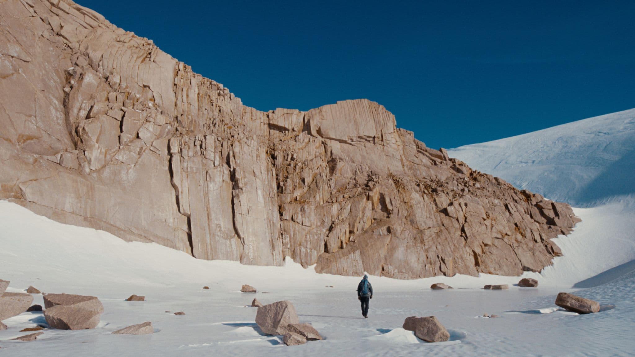 A lone hiker with a backpack crosses a snowy plain below a towering rock cliff in a polar landscape