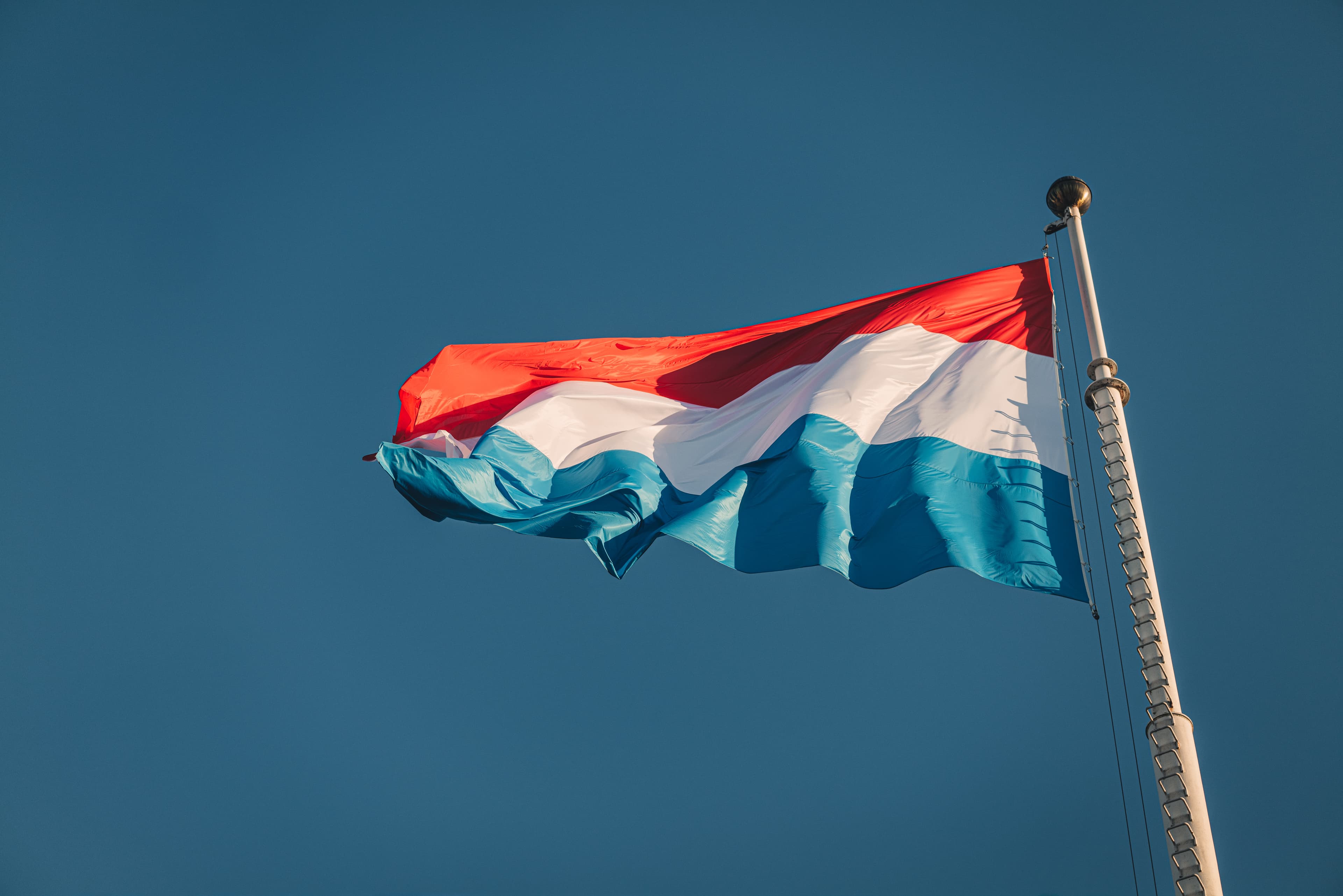 The red, white and light blue flag of Luxembourg waving on a pole against a blue sky